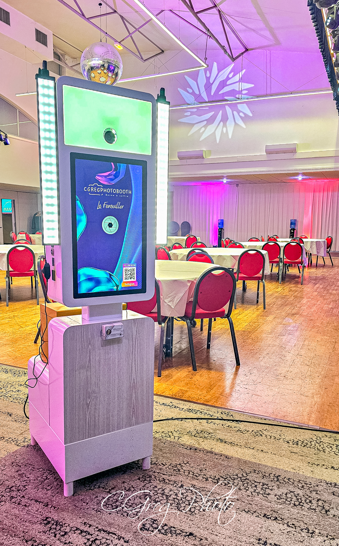 Au premier plan, la borne photo CGregPhotoBooth est allumée et prête à l'emploi. Derrière elle, la grande salle de réception s'organise doucement : les tables sont dressées, les chaises rouges alignées, et une douce lumière violette et rose commence à baigner l'espace, annonçant une soirée festive.