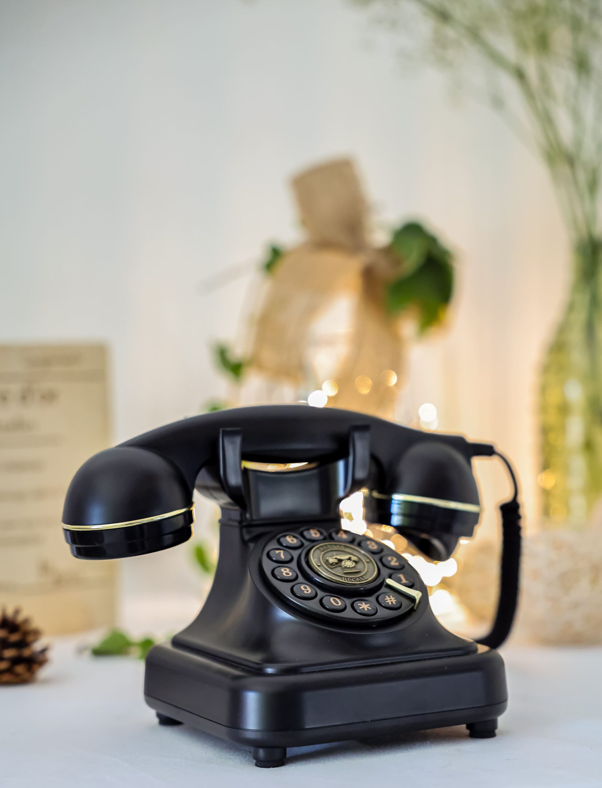 Sur une surface blanche, un téléphone rétro noir avec des liserés dorés capte l'attention. En fond, légèrement flou, une guirlande lumineuse scintille parmi quelques feuillages, ce qui donne une ambiance douce et chaleureuse.